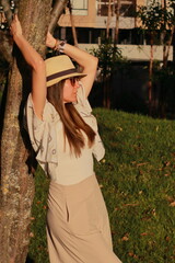 Obraz premium Young stylish woman wearing straw hat enjoying golden hour sunlight in a park.