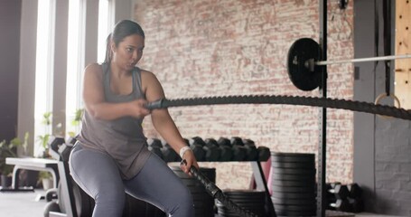 Entering gym and squatting before battle ropes, Asian woman alternating powerful waves for strength - Powered by Adobe