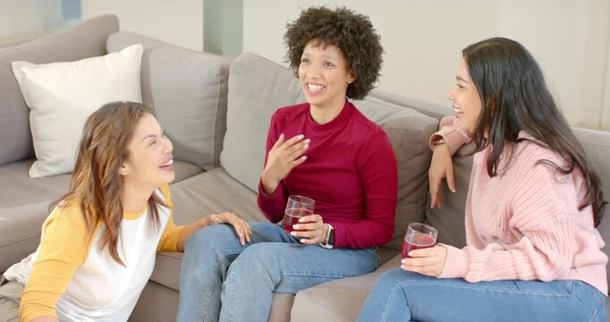 Diverse female friends sitting on sofa teasing snack recounting story and laughing for fun at home