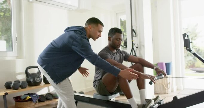Male trainer entering home gym guiding African American client on rowing machine for proper form