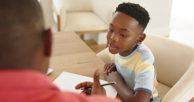 Father and son gesturing pencil toward notebook motivating son writing at table window, copy space