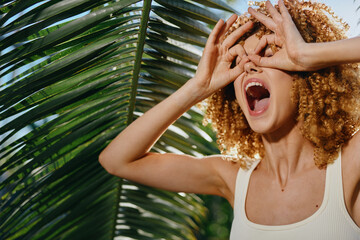 young woman with curly hair playfully making glasses gesture near green tropical palm leaves in...