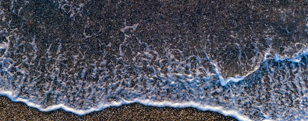 Top down view of white sea foam washing over dark sand and pebbles on a beach shoreline creating a natural texture pattern that represents the movement of the ocean tides and calm energy © Buğra Kaan Ersoy