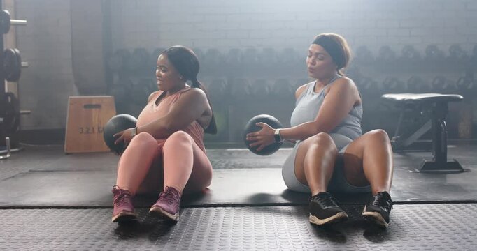 Diverse female friends starting workout picking up 3 kg medicine ball twisting torsos on gym floor