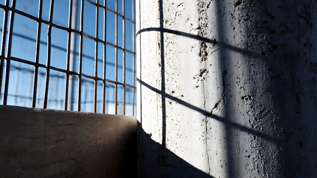 A concrete pillar adjacent to metal mesh fence creating layered grid-shadow shapes in strong sunlight