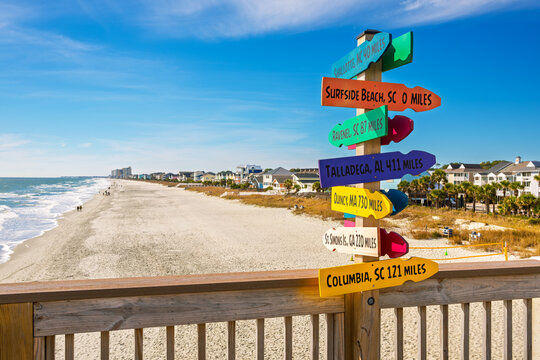 A colorful multi-directional signpost along the boardwalk and pier with the sandy Grand Strand beach coastline behind, in the resort town of Surfside Beach, South Carolina. - Powered by Adobe
