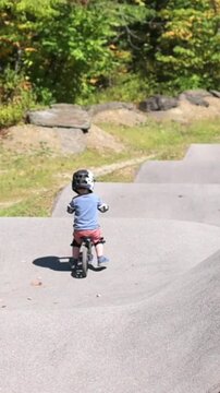 Verti al screen White toddler boy riding balance bike on pump track in Johnson City park usa, wearing helmet and knee pads, navigating asphalt bumps and small hills with focused