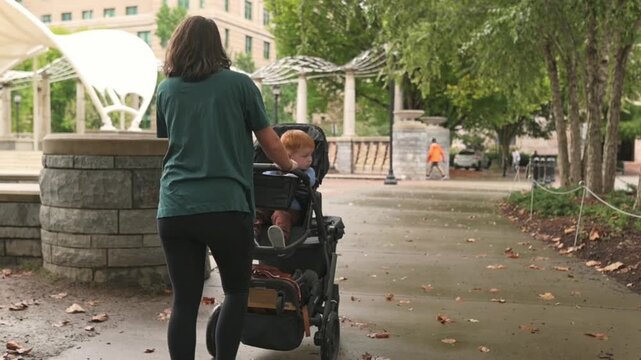 Woman surveys surroundings while guiding stroller, Female parent observes environment with caring oversight, Woman gently glances behind as she pushes stroller through park scenery