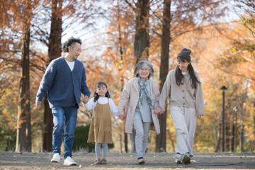 Fototapeta premium Autumn Family Photo: Three Generations Smiling in a Forest of Autumn Leaves Grandmother walking hand-in-hand with her grandchild and the child's married couple