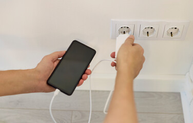 Charger connects smartphone phone via charging cable. Hands hold the adapter by a wall socket,...