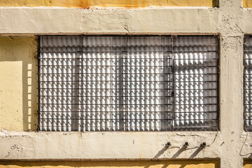 A building facade with a window grille casting shadows, pipes, and aged yellow wall, showing architectural texture and light interplay.