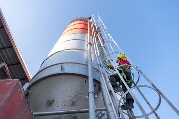 A low-angle shot of a construction worker in a safety harness climbing a tall cement silo ladder against a clear blue sky, emphasizing safety protocols.