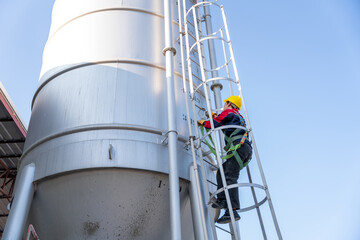 Close-up, low-angle shot of a construction worker in a safety harness climbing a cement silo ladder, looking down and ensuring safety.