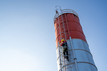 A construction worker in full safety gear climbs the fixed ladder of a tall, red and white cement silo under a clear blue sky.