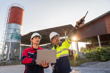 Two construction engineers reviewing factory operations with a laptop and radio at a concrete or cement plant. Safety and industrial teamwork concept.