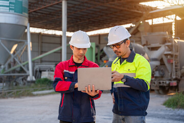Two engineers using a laptop for digital planning and quality control at a small concrete batching plant with mixer trucks in the background.