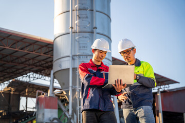 Two construction engineers working on a laptop near a large industrial silo at a cement or concrete production facility. Industrial technology and planning concept.
