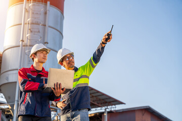 Two construction managers use a laptop and walkie-talkie for digital planning and site inspection, pointing toward the cement silo structure.
