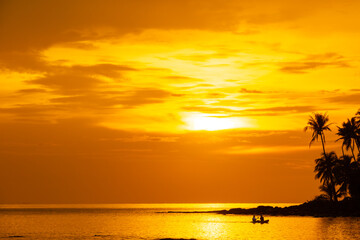 Nature tropical sunset paints the sky in fiery oranges and golds, with dramatic clouds. Silhouette palm trees and rocky coastline frame the setting sun as it dips towards the sparkling ocean horizon