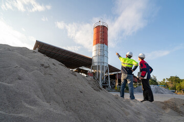 Two engineers inspecting a small concrete batching plant and aggregate piles at sunset. Focus on construction industry, ready-mix business, and small-scale operations.