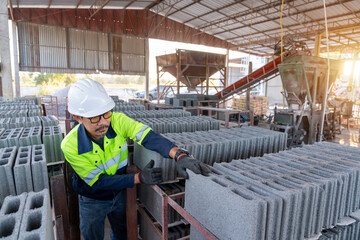 A construction manager inspects the quality of fresh concrete cinder blocks with the production line and machinery visible in the background.