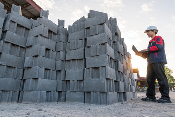 A construction manager uses a laptop to manage inventory and review data, standing next to a large stack of fresh concrete cinder blocks.