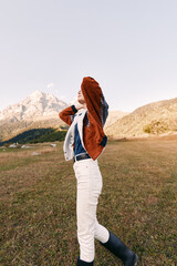 Obraz premium Woman in mountain meadow wearing autumn fashion jacket and boots, portrait of relaxed style outdoors. White pants, knit sweater and scenic landscape for travel and lifestyle.