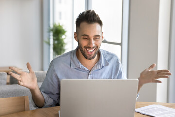 Happy Hispanic man sits at table looking at laptop screen and gesturing during videoconference event, reacting enthusiastically on good news in e-mail or notifice, celebrating results, feels excited
