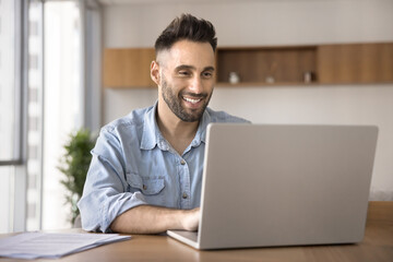 Hispanic man working on laptop sits at table at home office, smiling, reading good news in email,...