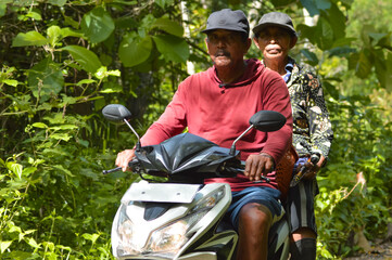 A man in a red shirt and a woman in a patterned black and gold top pause on a white scooter along a rural path surrounded by dense tropical foliage, looking directly at the camera. © agratitudesign