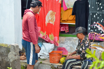 A man stands observing while a woman sits on a concrete step outside a home. She removes papaya and banana leaves from an orange basket, representing their fresh harvest from the fields.