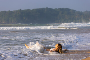 Fototapeta premium Young Woman Lying on the Shoreline as Waves Wash Over Her at Talalla Beach, Sri Lanka