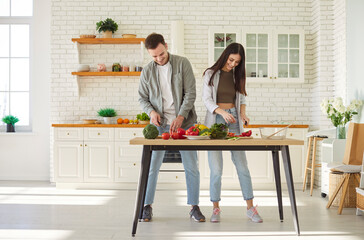 Happy young couple man and woman cooking in modern kitchen cutting fresh vegetables preparing healthy vegan salad at home together. Diet, vegetarian food, healthy lifestyle and nutrition concept.