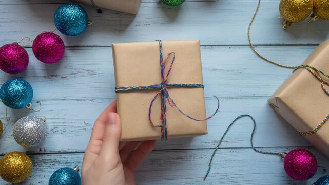 A hand holding a beautifully wrapped Christmas gift box surrounded by colorful festive ornaments on a wooden background.