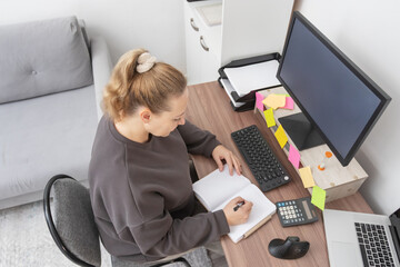 Caucasian woman working from home, planning and writing notes at a wooden desk with a computer monitor, showcasing a casual remote office setup.