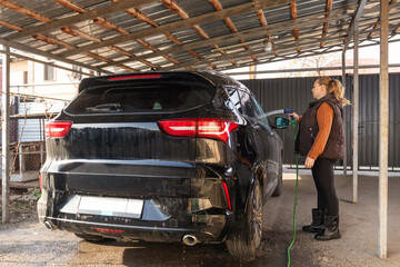 Middle-aged woman washing her black SUV vehicle under a carport at home. Female doing auto maintenance and care routine using a water hose.