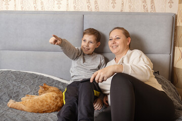 Happy Caucasian mother and preteen son relaxing together at home, sitting on the bed and watching television with a remote control.