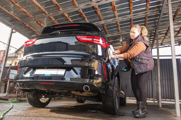 Middle-aged woman washing her black SUV vehicle under a carport at home. Female doing auto maintenance and care routine using a water hose.
