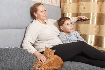 Happy Caucasian mother and preteen son relaxing together at home, sitting on the bed and watching television with a remote control.