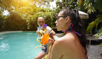 A man and a woman sit by the pool with cocktails and fruit.