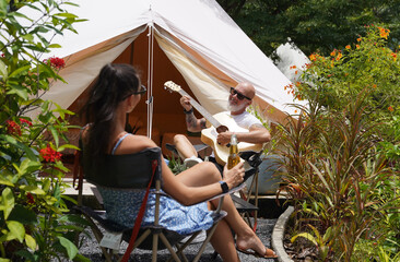A man plays guitar for a woman near a cozy tent at a glamping site.