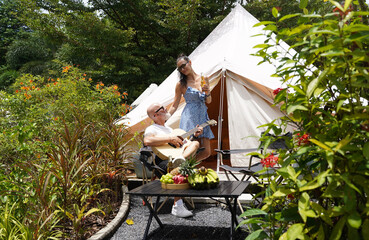 A man plays guitar for a woman near a cozy tent at a glamping site.