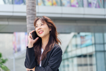 Fototapeta premium Young Asian business woman, busy with work on street, wearing black suit talking on phone with happy smile, enjoying modern urban cityscape, professional working outdoors