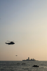 Trivandrum India December 3 2025 : Indian Navy helicopter and warship operate together at sunset off Shangumugham Beach with small boats in foreground during Navy Day demo. © Sooraj