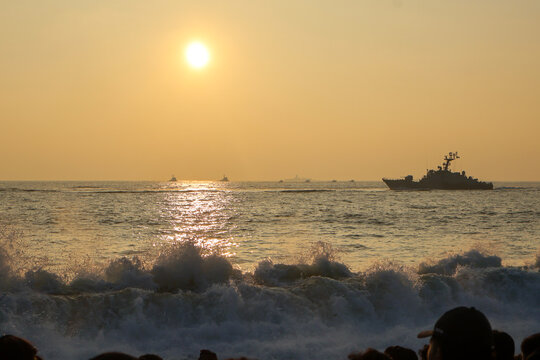 Trivandrum India December 3 2025 : Indian Navy warship powers past crashing waves at golden sunset while crowds watch from Shangumugham Beach during Navy Day demo. - Powered by Adobe