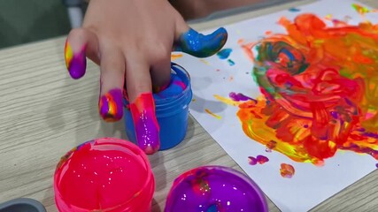 A child's hand with brightly colored fingers dips his finger into a jar of blue finger paint next to a colorful painting on the table.