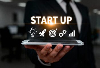 Businessman showing start up text with light bulb rocket target gear and growth graph icons hovering over a smartphone