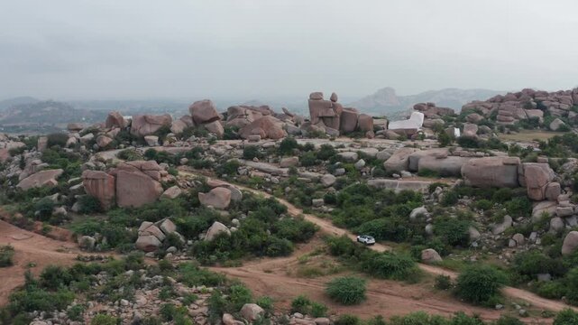 White SUV winds along a dusty trail through Hampi&rsquo;s dramatic granite boulder hills, captured in a wide aerial shot that highlights the rugged landscape and sense of cinematic adventure.