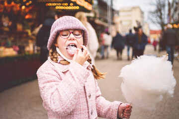 Little cute kid girl eats sweet cotton candy on Christmas market. Happy child on traditional Xmas...