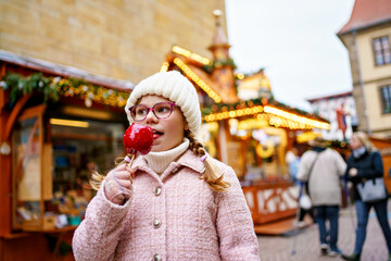 Obraz premium Little cute kid girl eat sweet sugar covered red candy apple on Christmas market. Happy child on traditional Xmas fair in Germany.